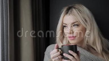 美丽的金发女人拿着陶瓷杯的茶或咖啡享受。 在床上喝茶。 看着窗户喝茶